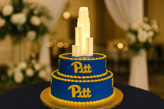 A three-tiered University of Pittsburgh groom’s cake decorated in Pitt’s blue and gold colors, topped with a glowing 3D printed LED figurine of the Cathedral of Learning, displayed at a wedding reception.