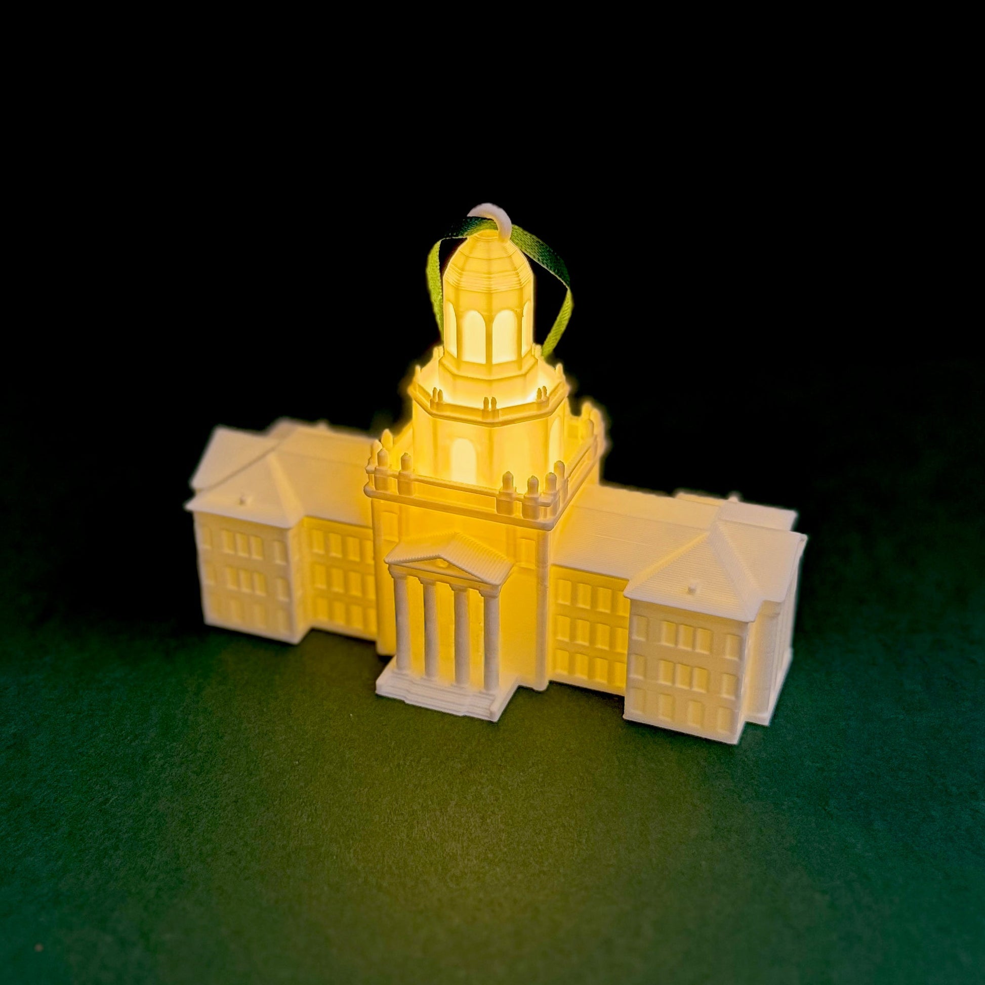 A glowing 3D-printed Pat Neff Hall Christmas ornament with a dark green hanging ribbon, resting on a dark green background.