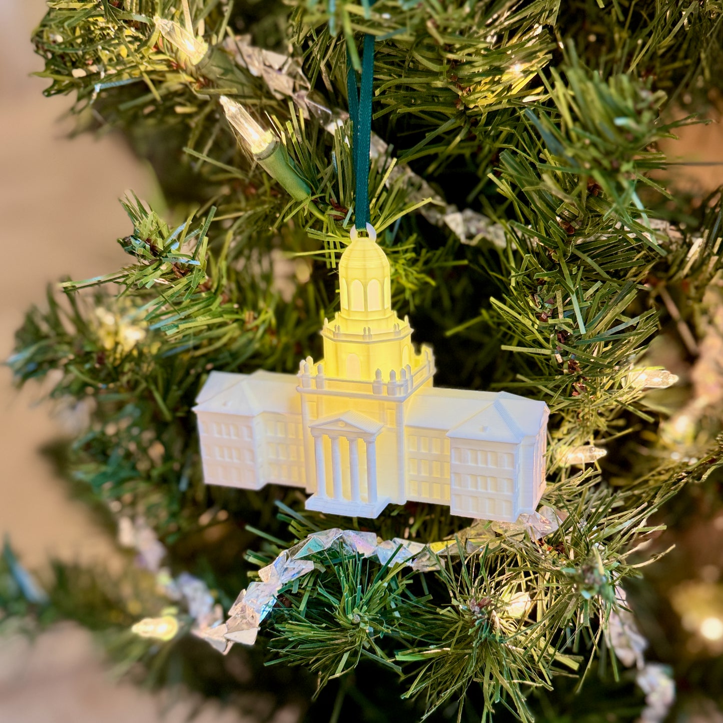 A lit Baylor University Pat Neff Hall ornament hanging on a Christmas tree surrounded by warm holiday lights.