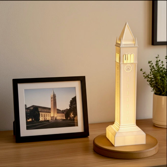 Illuminated 3D-printed model of Berkeley’s Campanile tower on a wooden base, placed beside a framed photo of the actual tower and a small potted plant on a desk.