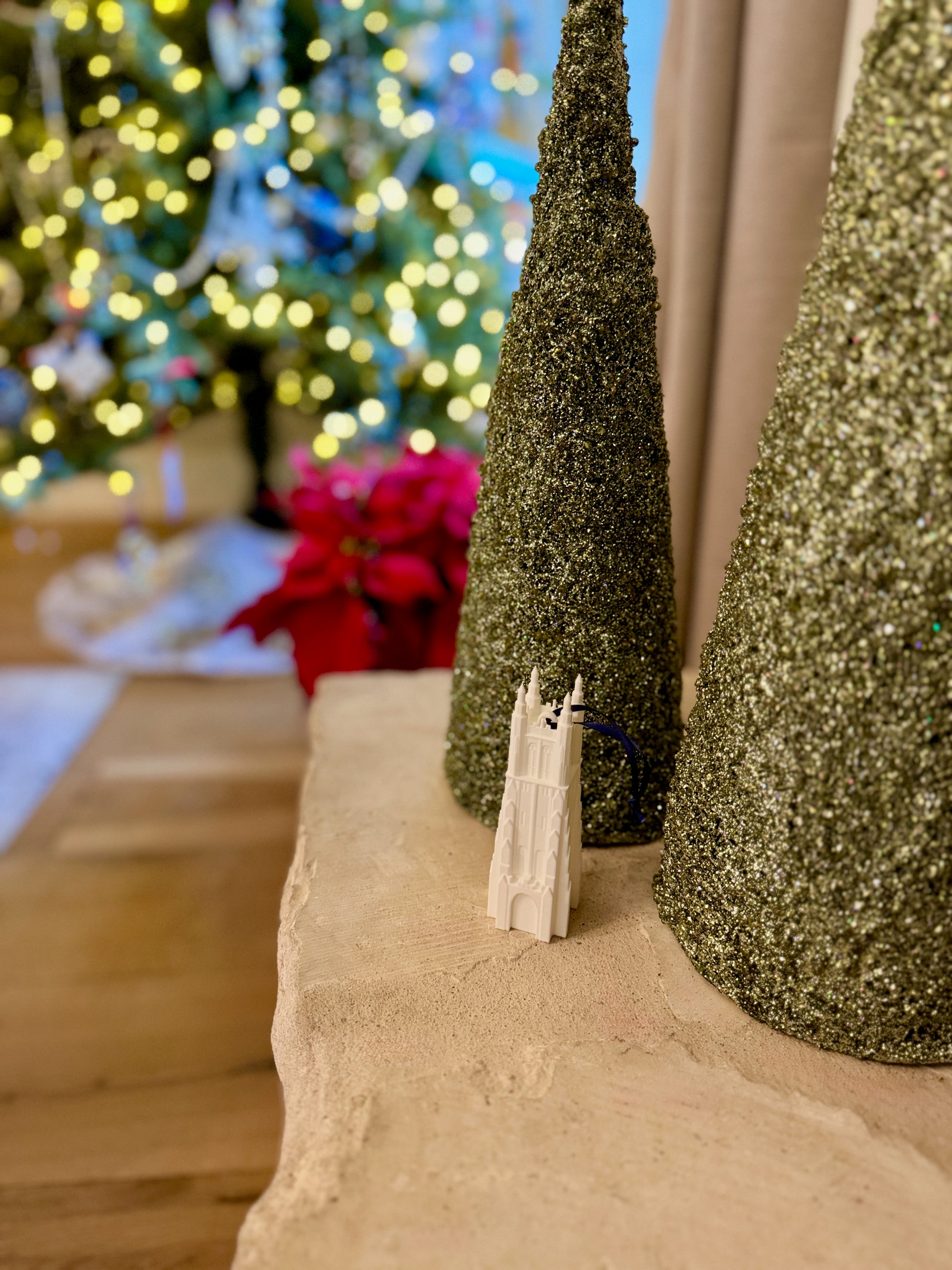 Decorative setup with glittery trees and a Duke Chapel ornament on a surface, with a blurred Christmas tree in the background.