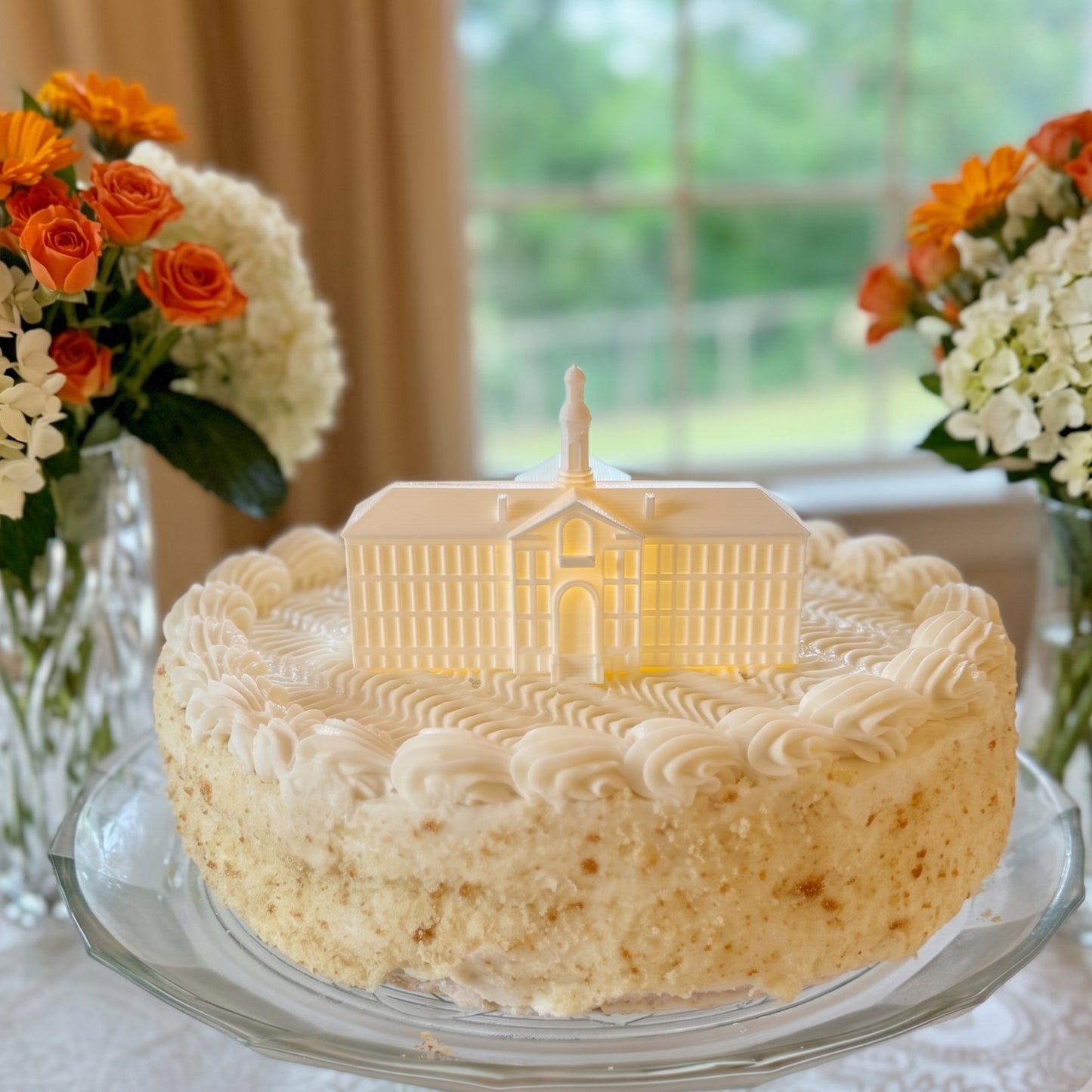 A frosted groom's cake on a glass pedestal, topped with a detailed, cake topper of the Princeton University Nassau Hall building, with bouquets of white hydrangeas and orange flowers in the background.