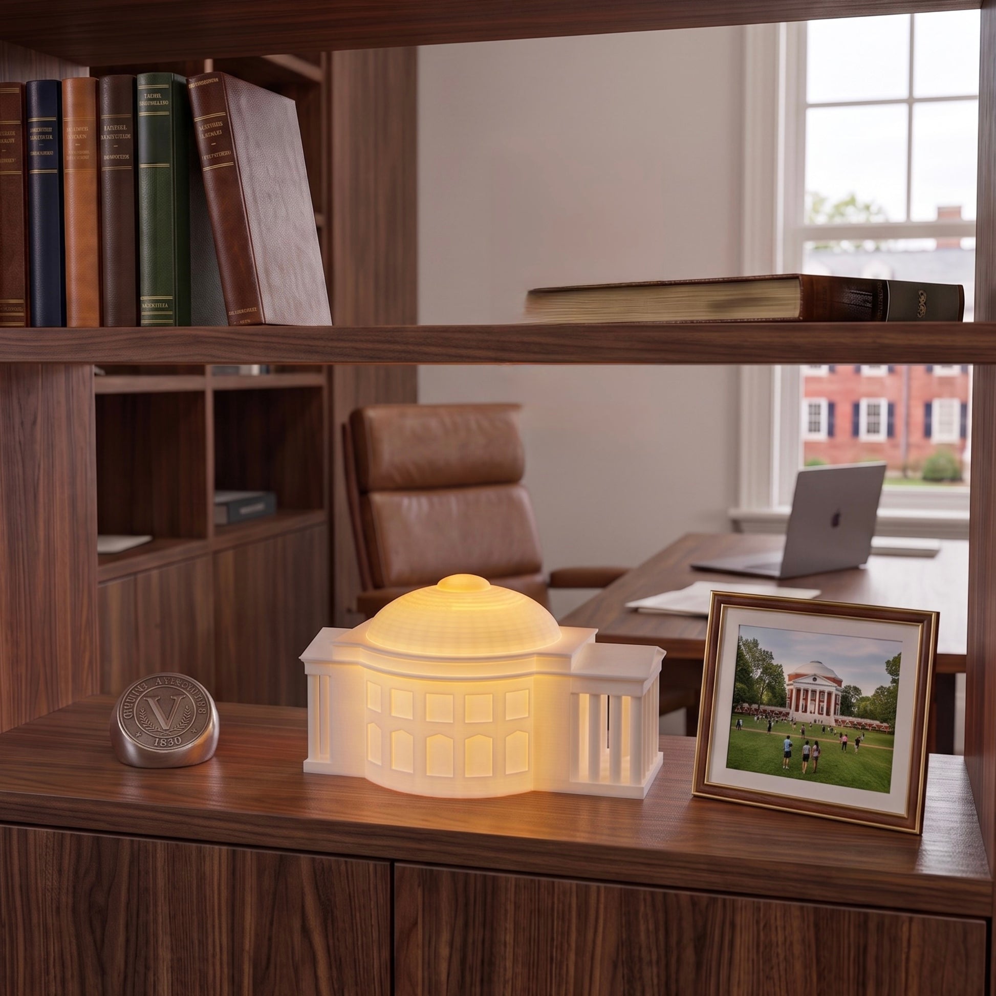 A miniature illuminated replica lamp of the UVA Rotunda, glowing warm white on a wooden shelf in an office of a UVA Alumni, next to a framed photo of the University of Virginia campus.
