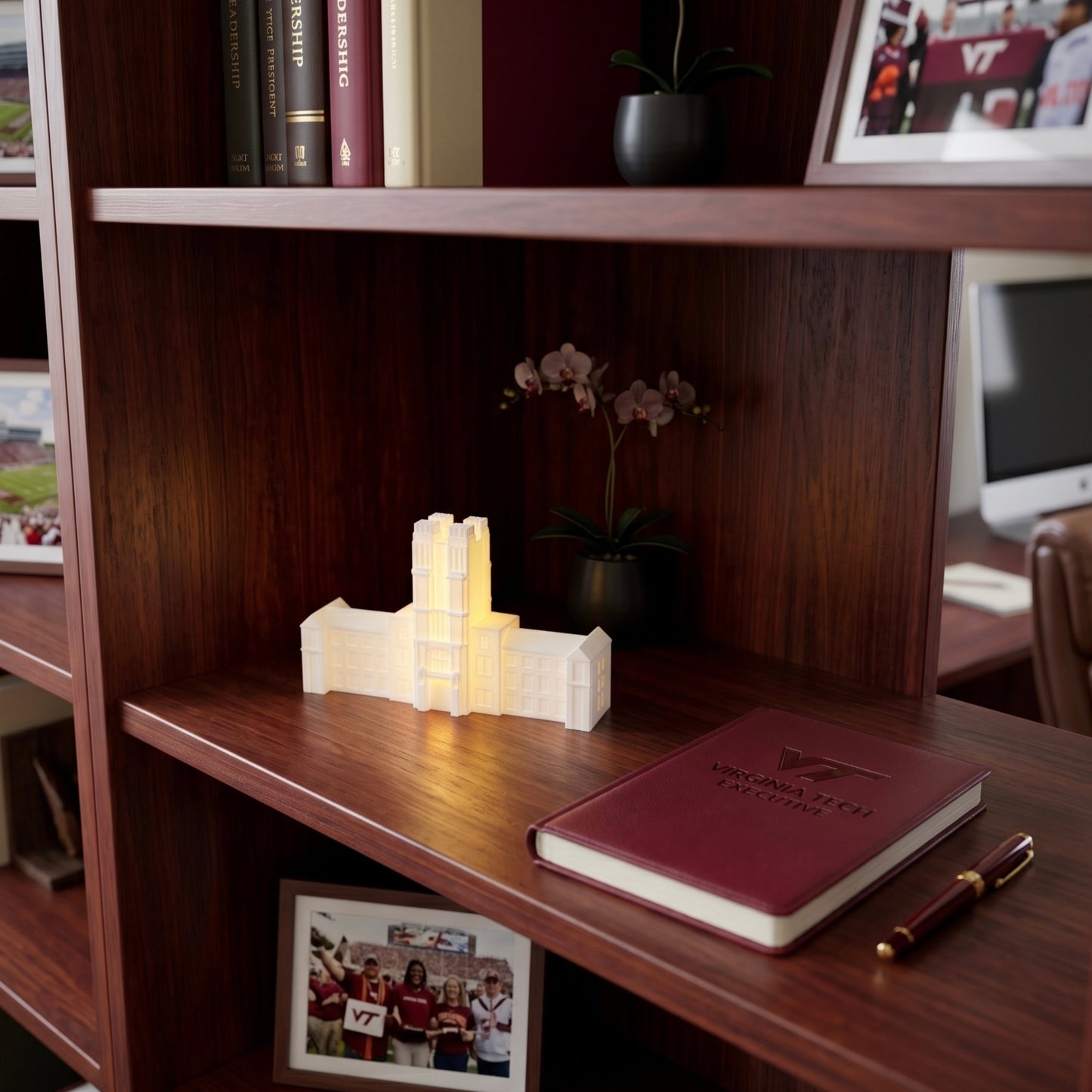 A glowing white replica of a Virginia Tech building sitting on a dark wood office bookshelf next to a maroon notebook and pen.