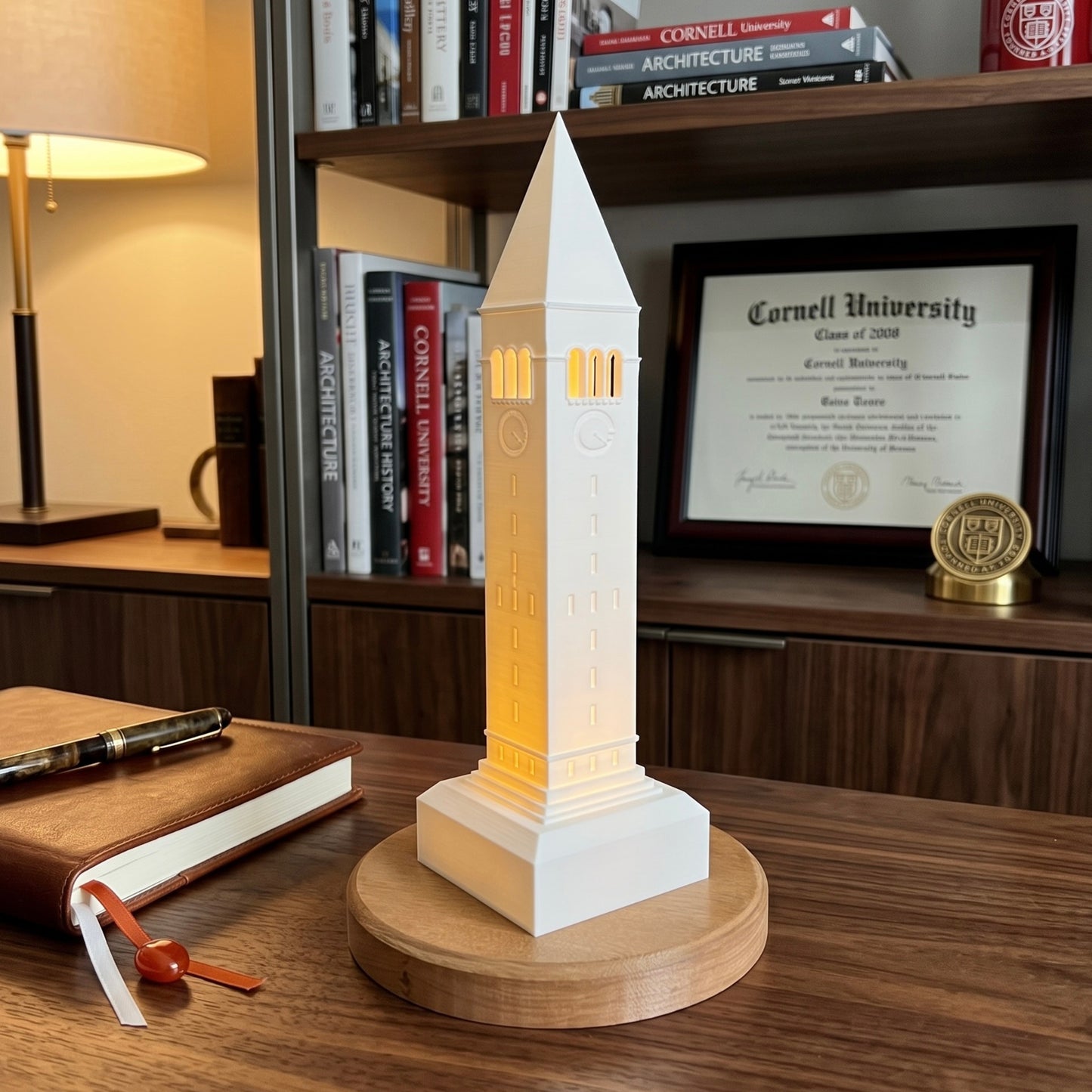 A Cornell University-themed illuminated model tower lamp on a round wooden base, set on a wooden desk in front of a bookshelf with books, a lamp, and a framed Cornell University diploma.